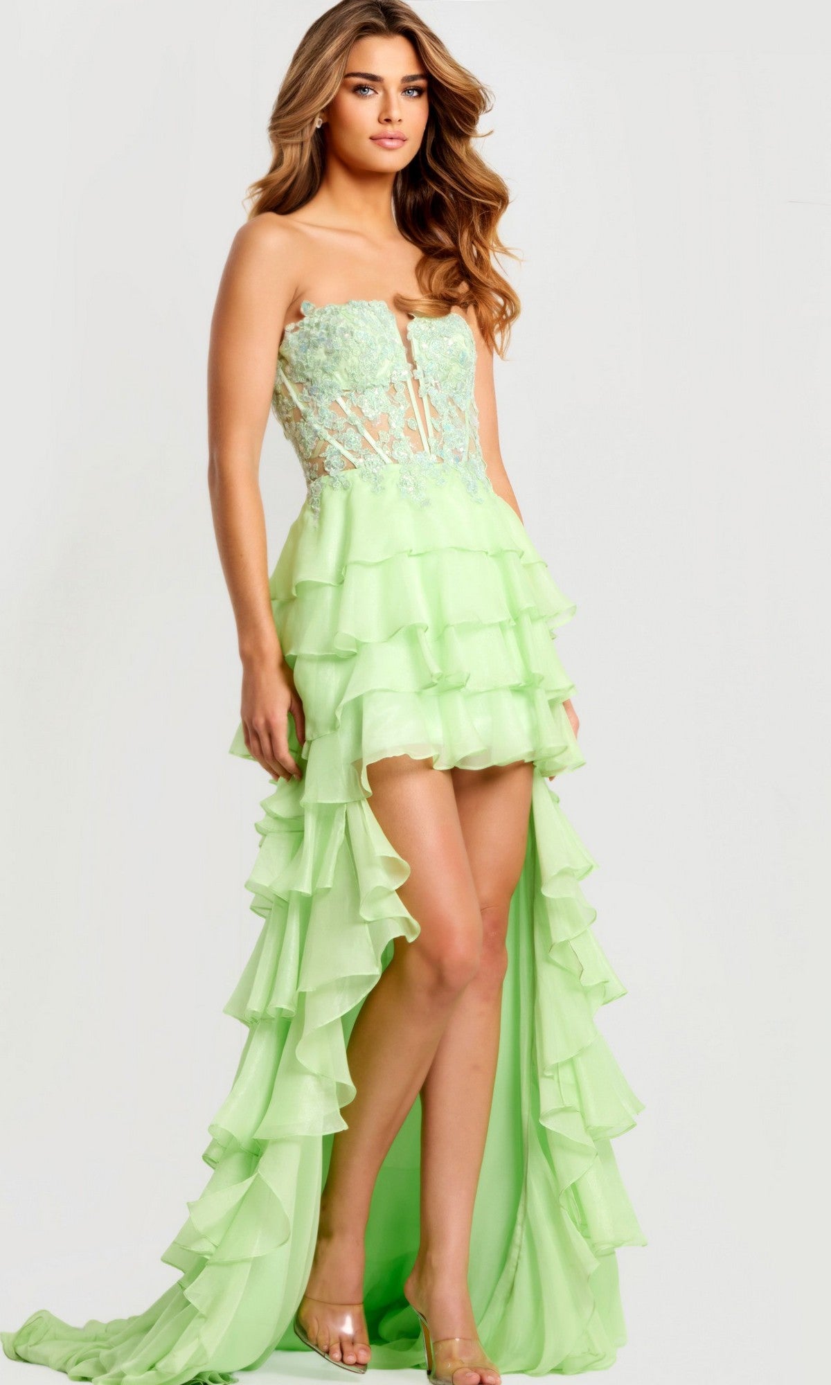 A woman wears the Jovani 47422 long prom dress in light green, featuring a strapless lace-embroidered corset bodice and a tiered high-low chiffon ruffle skirt that's short in front and long at the back.